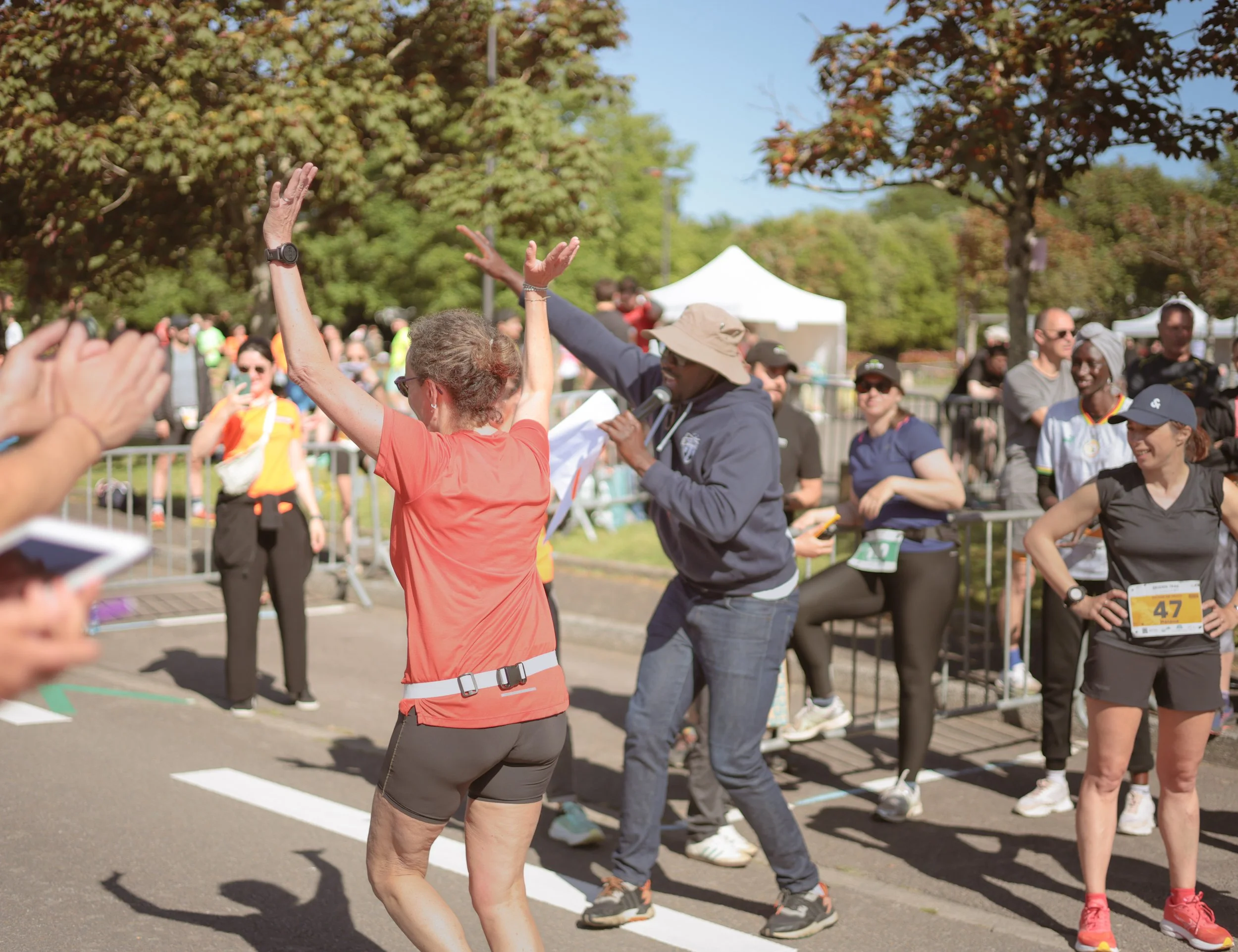 Une femme avec une robe orange et une montre au poignet se tient avec les bras levés, tandis qu'un homme, avec un chapeau beige, une veste grise et un micro, semble l'encourager. Autour d'eux, plusieurs personnes en tenue de course, portant des numéros, participent à un événement de course en extérieur, avec des arbres et des tentes en arrière-plan.