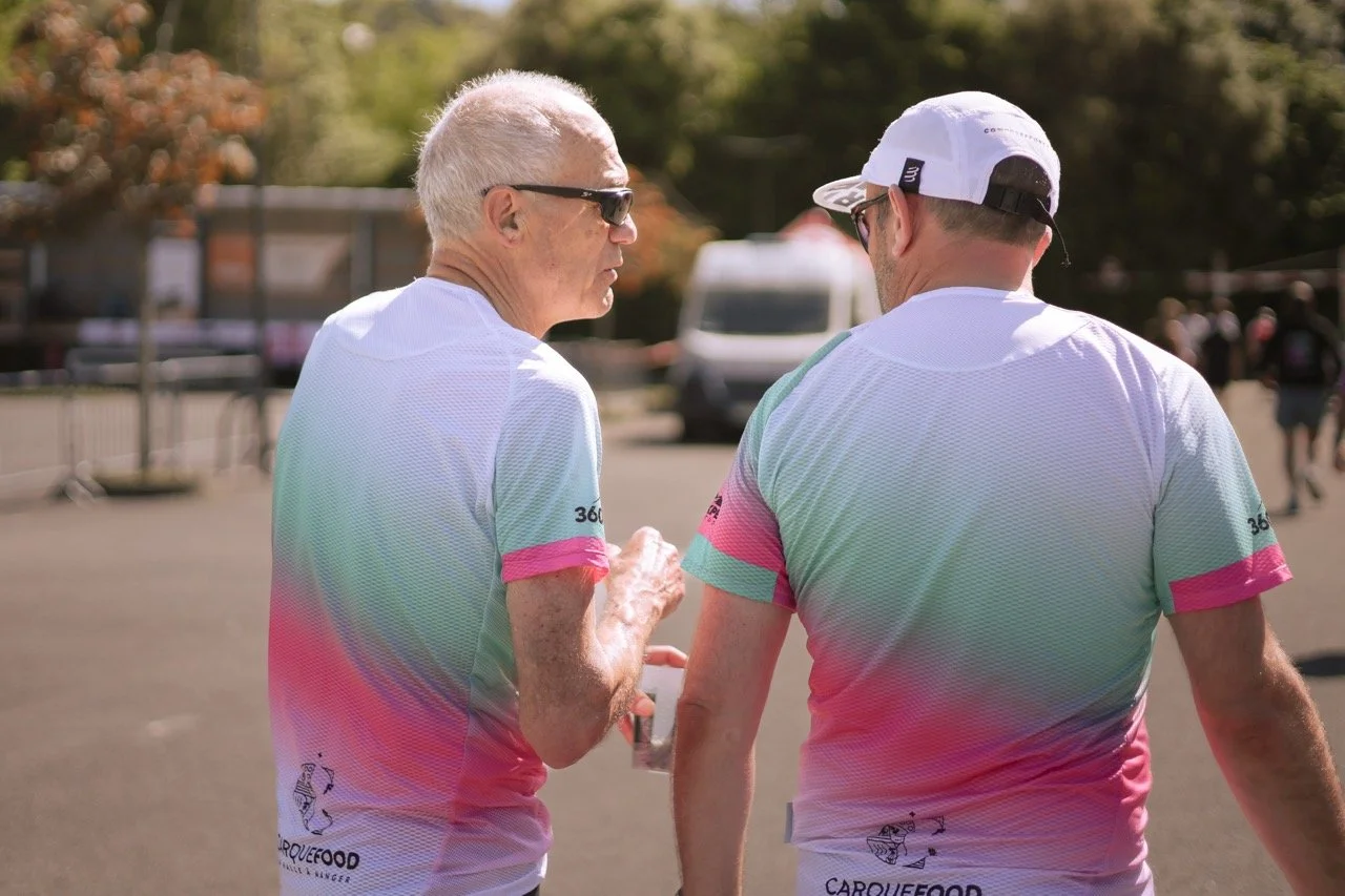 Deux hommes portant des t-shirts blancs avec des dégradés de couleurs pastel, discutant lors d'un événement en extérieur.