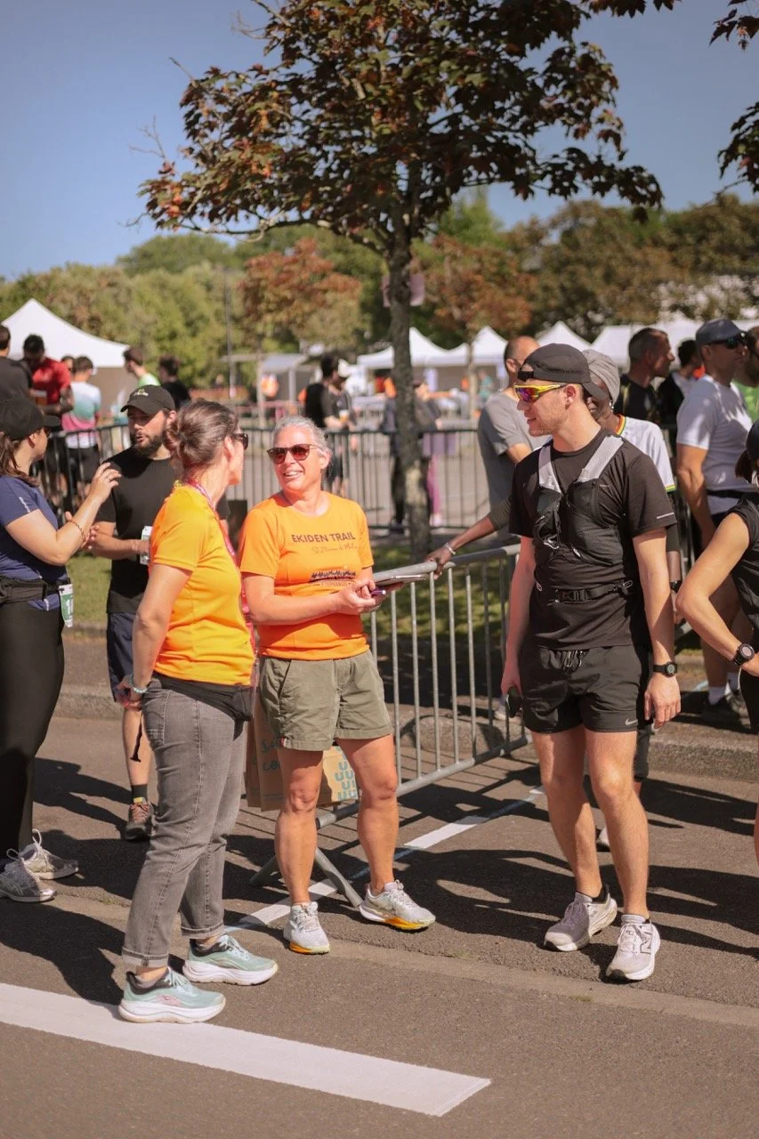 Groupe de personnes à une course ou un événement sportif, en plein air, avec des tentes et des barrières de sécurité, certains portant des vêtements de course, d'autres discutant.