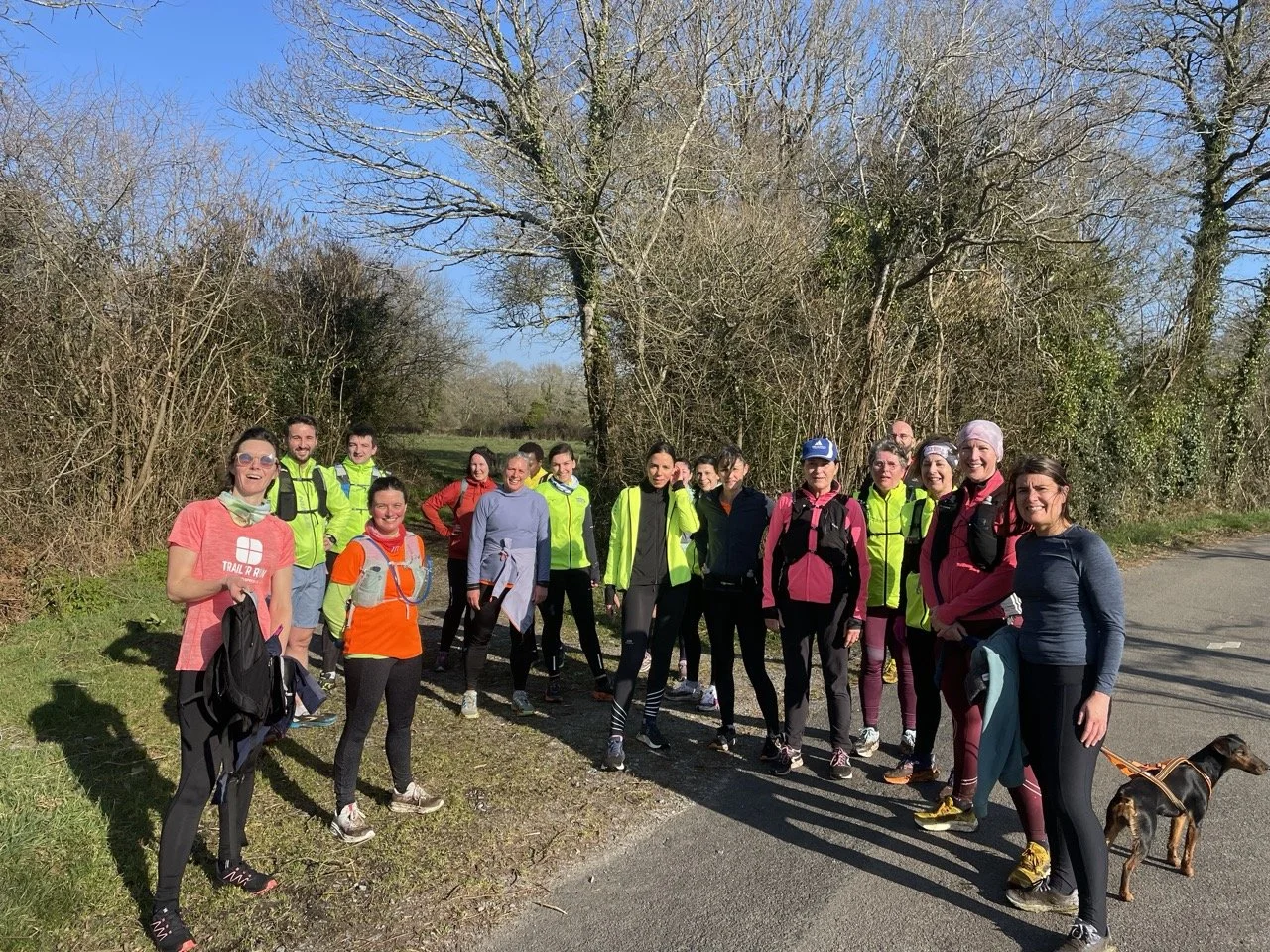 Groupe de personnes en tenue de course à pied, posant dans la nature avec des arbres sans feuilles, sous un ciel bleu, accompagnés d'un chien.