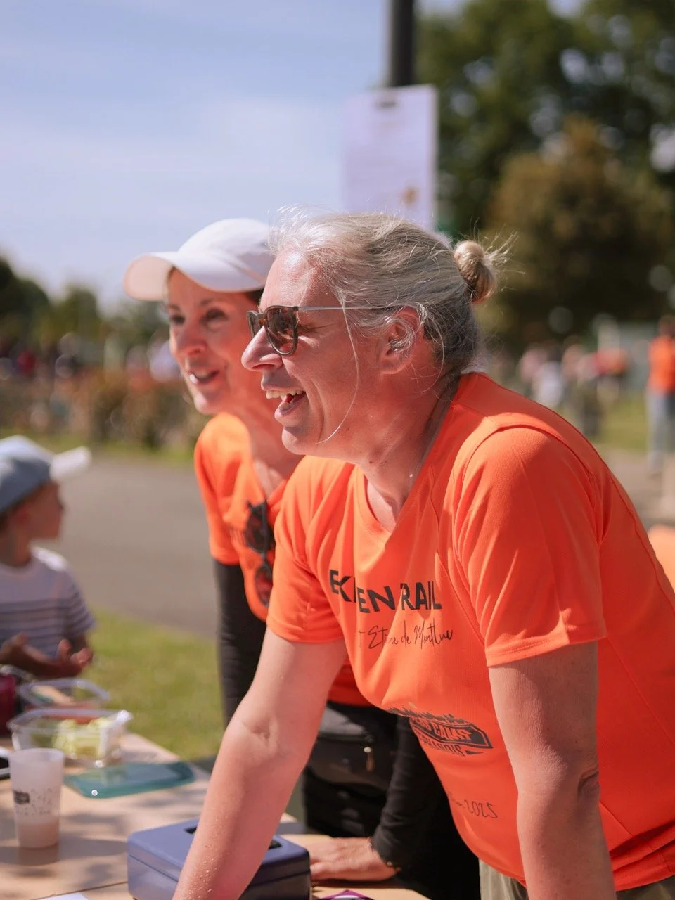 Deux femmes âgées souriantes portant des t-shirts orange à un événement en plein air, probablement une course ou une marche, avec des personnes en arrière-plan.