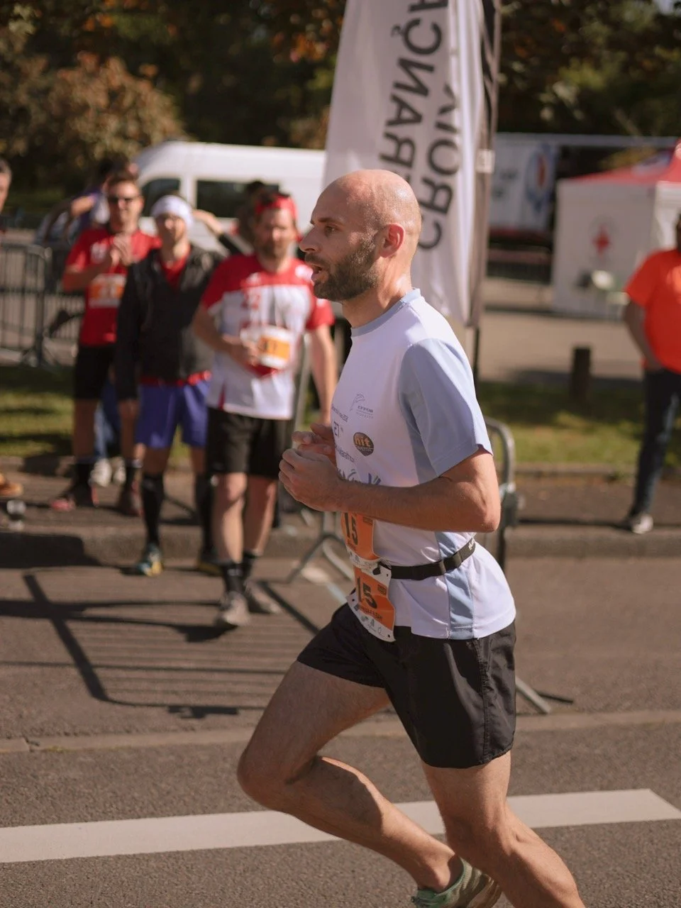 Un homme court lors d'une course, portant un t-shirt blanc et un numéro de bib orange, avec un groupe de spectateurs ou autres coureurs en arrière-plan.