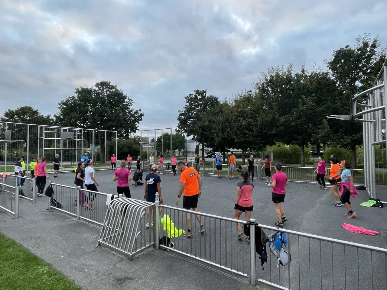 Groupe de personnes faisant une séance d'exercice en extérieur dans une cour clôturée, avec des vêtements de sport colorés, sous un ciel nuageux, entouré d'arbres.