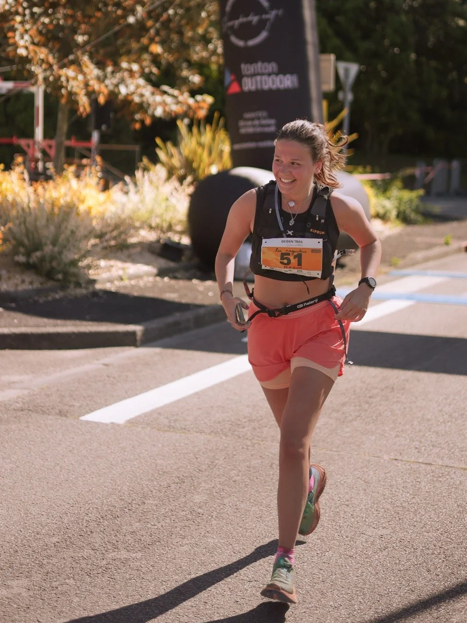 Une femme courait lors d'une course en plein air, portant un dossard numéro 51, un débardeur noir, un short rose, et une montre. Elle sourit en courant sur une route ensoleillée.