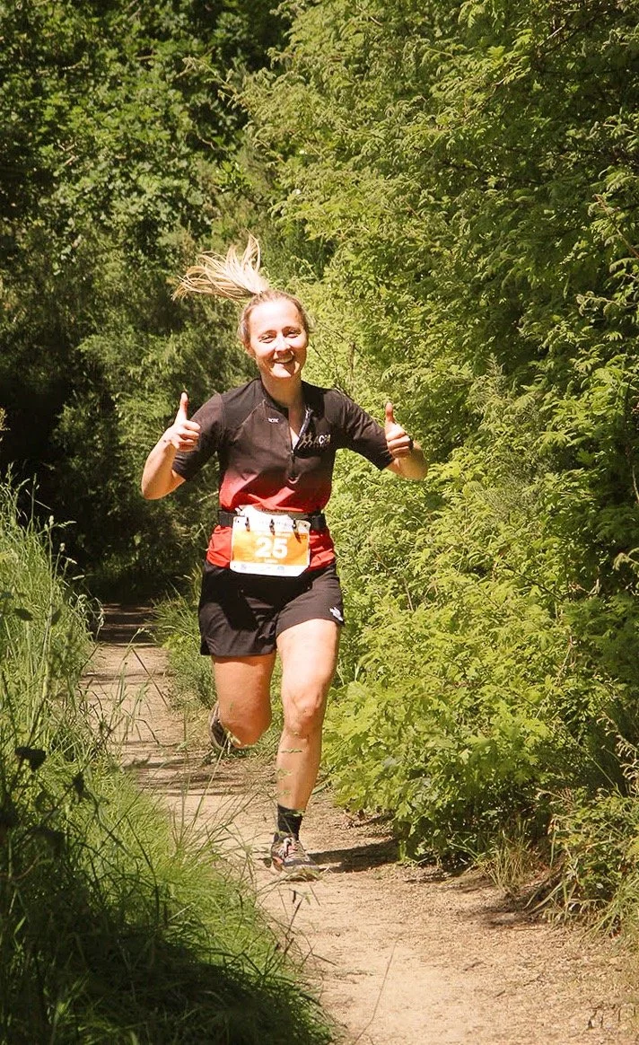 Une femme court sur un sentier en forêt, souriant et faisant un signe de pouce en l'air.