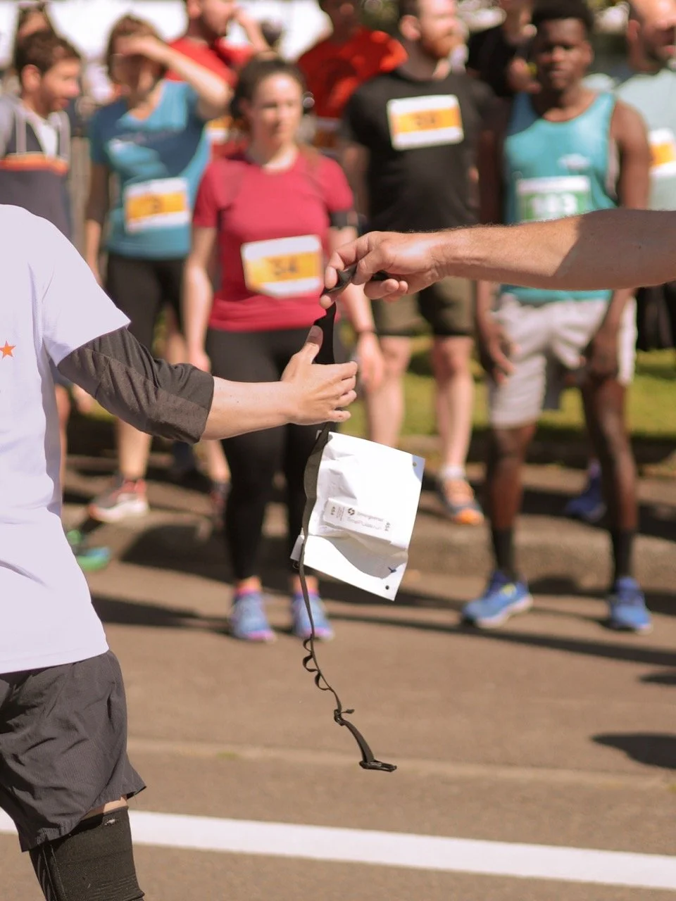 Une personne remet un dossard à une autre lors d'un événement de course à pied, avec d'autres participants en arrière-plan.