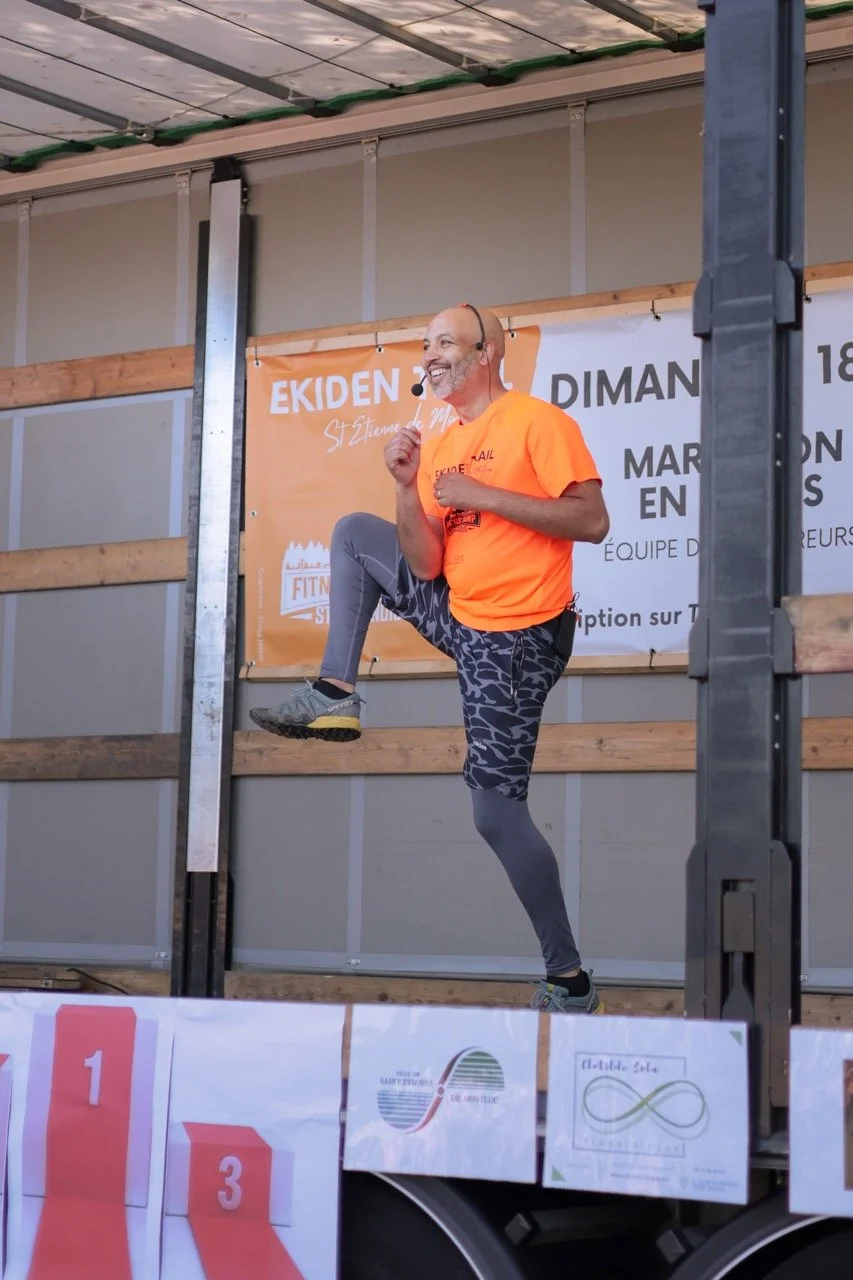 Un homme souriant en tenue de sport orange et pantalon gris à motifs, levant un genou en l'air lors d'une compétition de course à pied sur une structure en bois et métal.