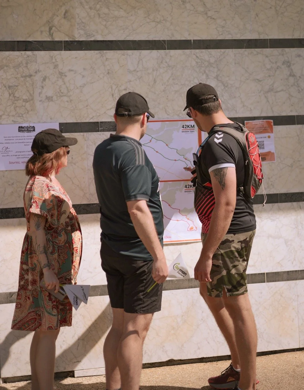 Trois personnes regardent une carte affichée sur un mur en marbre. Deux hommes en vêtements décontractés avec casquettes, et une femme en robe colorée et casquette.
