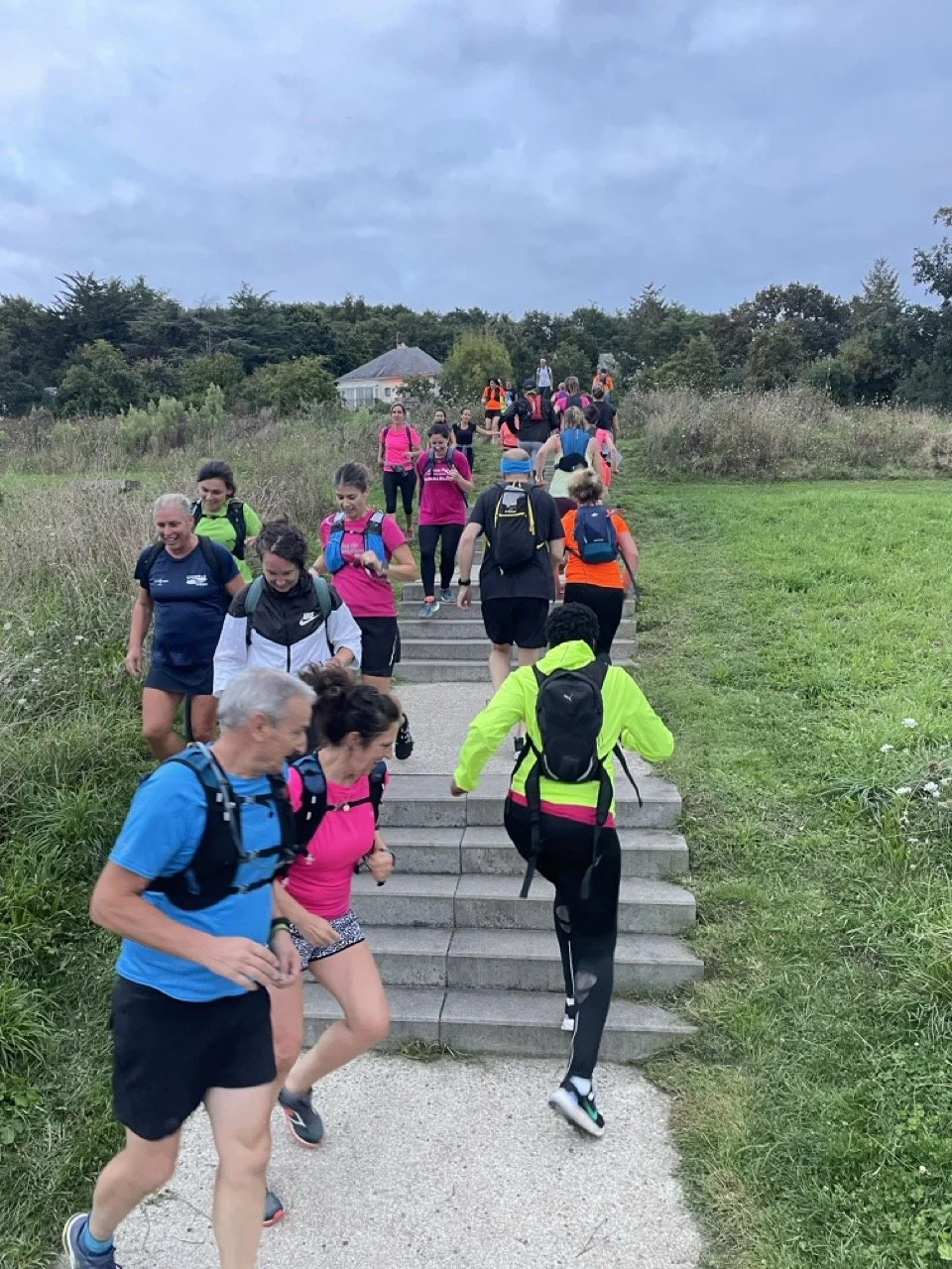 Groupe de personnes faisant de la marche ou de la course en montée sur un sentier avec des escaliers en extérieur, sous un ciel nuageux.
