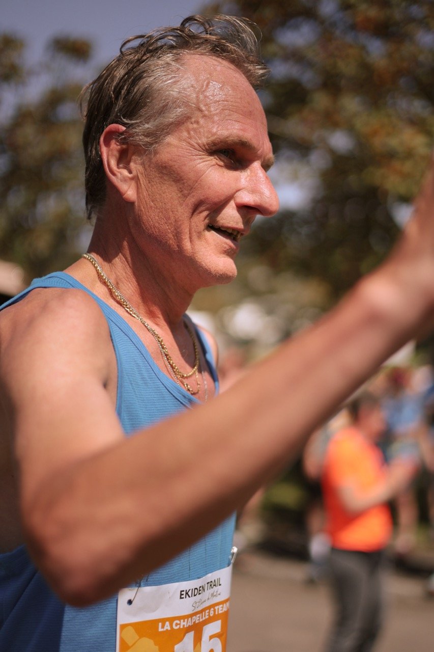 Un homme âgé souriant, portant un maillot de course bleu, avec un dossard orange, participe à une course en plein air, sous un ciel ensoleillé.