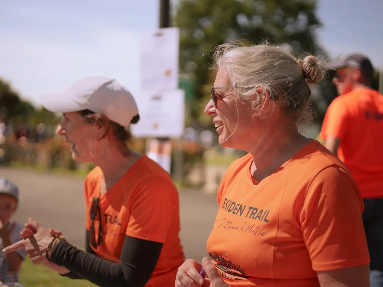 Deux femmes souriantes en chemises orange participant à une course ou un événement en plein air, avec un fond flou de personnes et de nature.