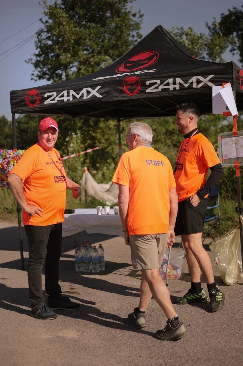 Trois hommes vêtus de t-shirts orange, dont deux portent des casquettes, discutent sous une tente noire avec le logo '24MX'. Un homme a un t-shirt avec l'inscription 'STAFF'. Il y a plusieurs bouteilles d'eau sur le sol et une table avec des petits v