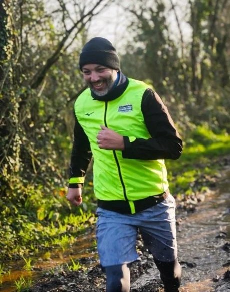 Homme souriant en veste jaune fluorescent et bonnet noir, courant dans la nature sur un sentier forestier.