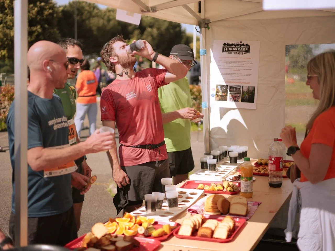 Groupe de coureurs en pause à un stand de ravitaillement lors d'une course, avec des sandwiches, des oranges et des boissons à leur disposition.