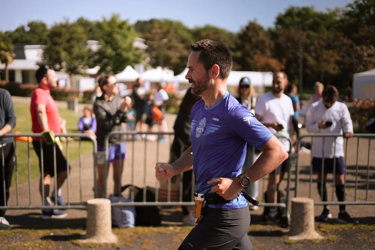 Un homme en t-shirt bleu court lors d'une course ou d'un marathon dans un parc, avec un groupe de spectateurs ou de participants en arrière-plan, entouré d'une barrière métallique.