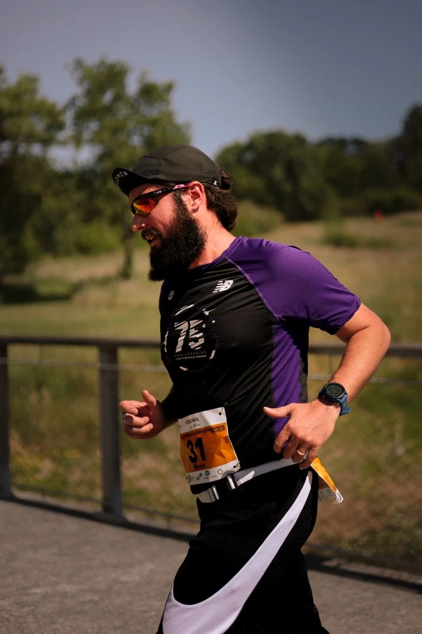 Un homme courrant lors d'une course en plein air, portant une maillot noir et violet, des lunettes de soleil et une montre sportive, avec un numéro de course 31 attaché à sa ceinture.