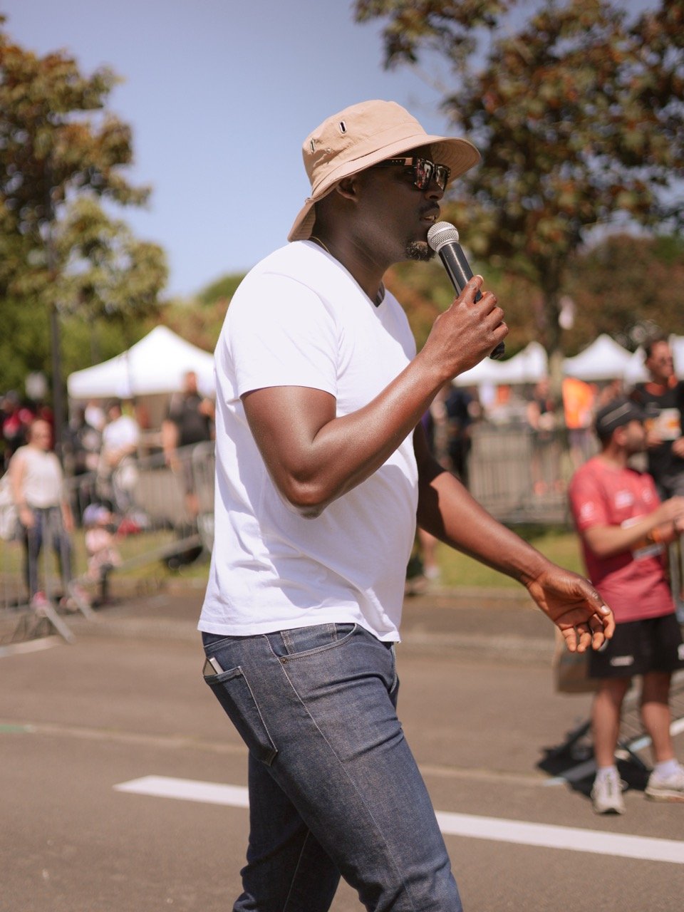 Homme portant un chapeau beige, lunettes de soleil, t-shirt blanc, tenant un microphone lors d'un événement en plein air with des personnes autour.
