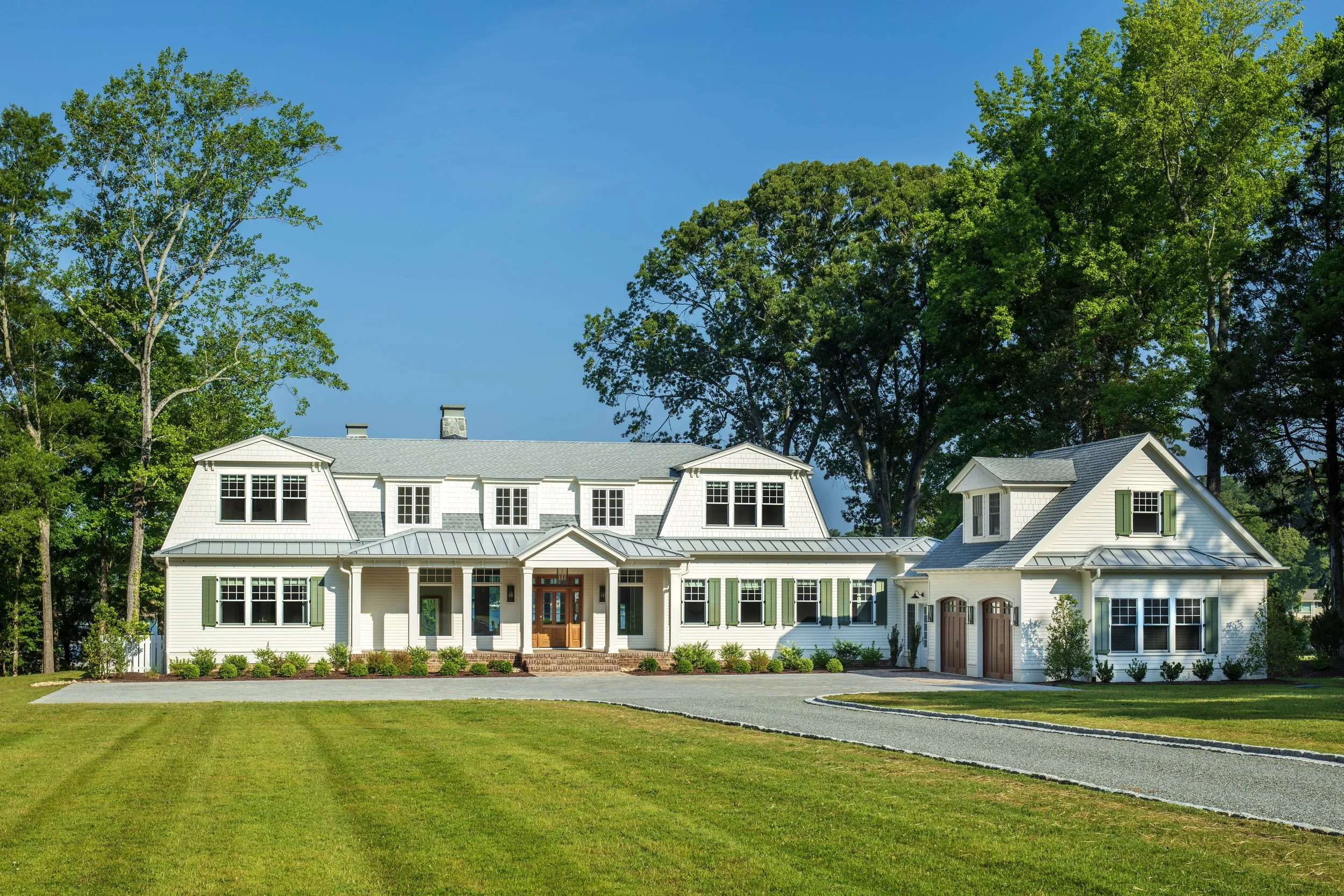 A large, coastal, white house with a gray gambrel roof and multiple windows, surrounded by green trees and a well-maintained lawn, with a paved driveway leading to the garages. Cape Cod inspired architecture on the Northern Neck in Virginia.