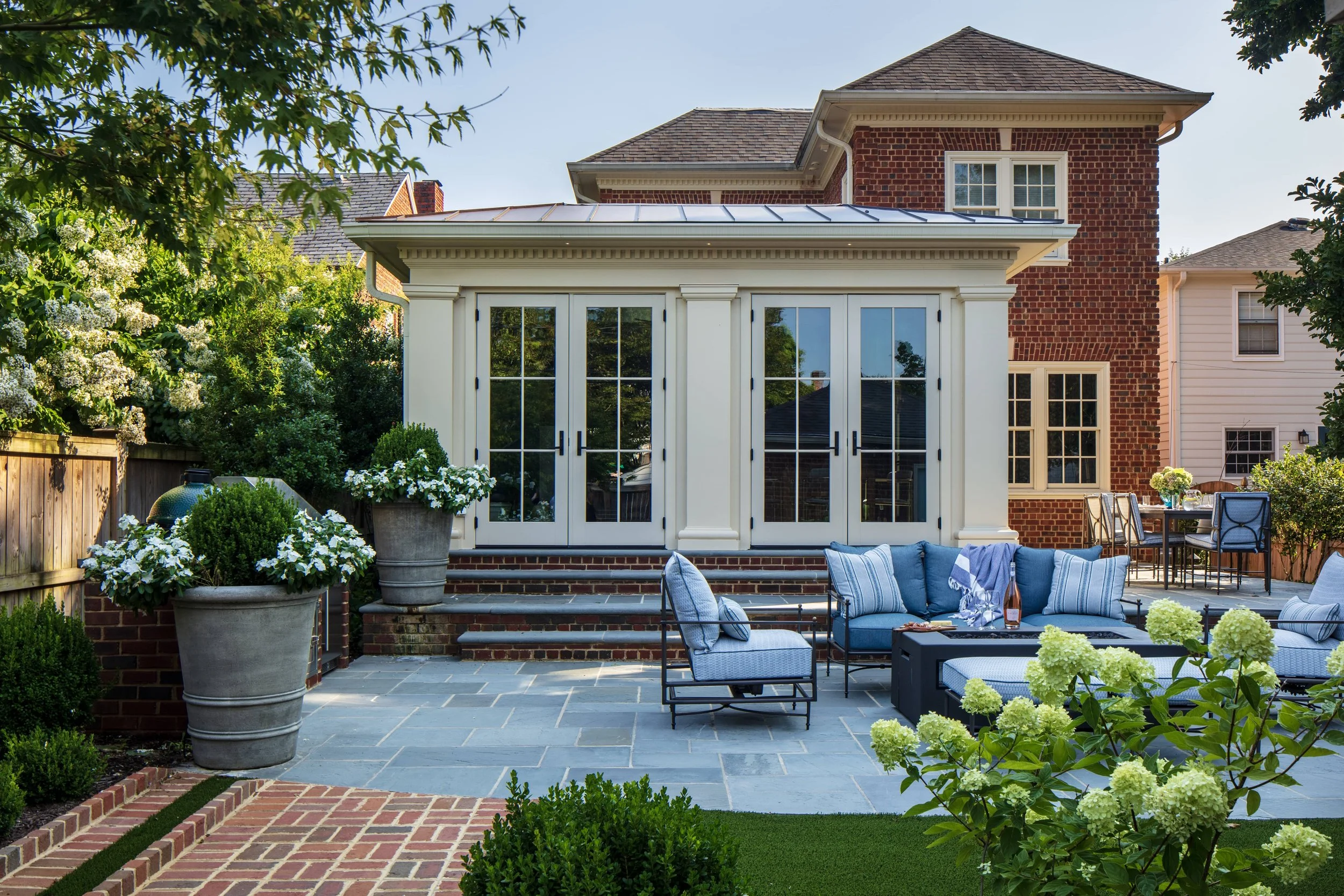 A timeless backyard patio with blue outdoor furniture, potted plants, and a screened porch attached to a brick house with large glass doors in Richmond, VA.