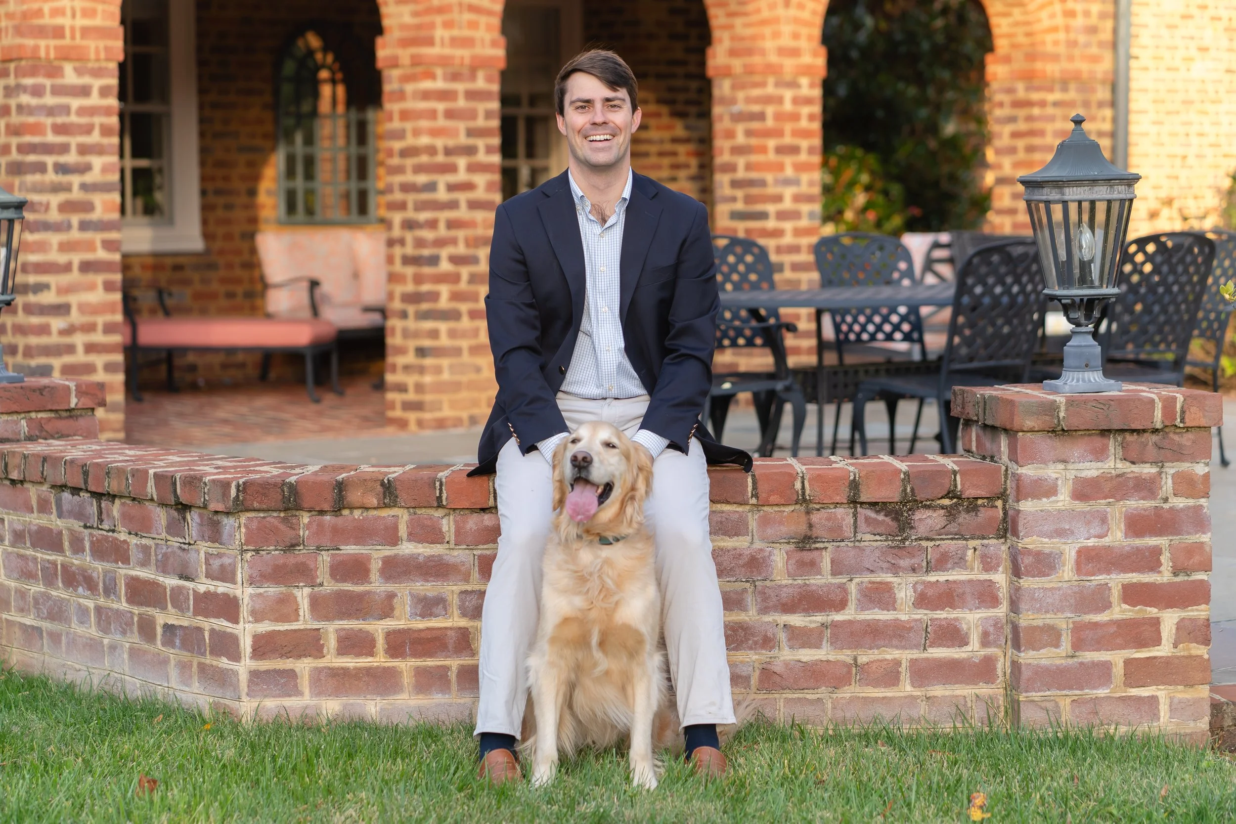 A man in a dark blazer and light-colored pants sitting on a brick wall with a happy golden retriever in front of him, outdoors in front of a brick building with patio furniture.