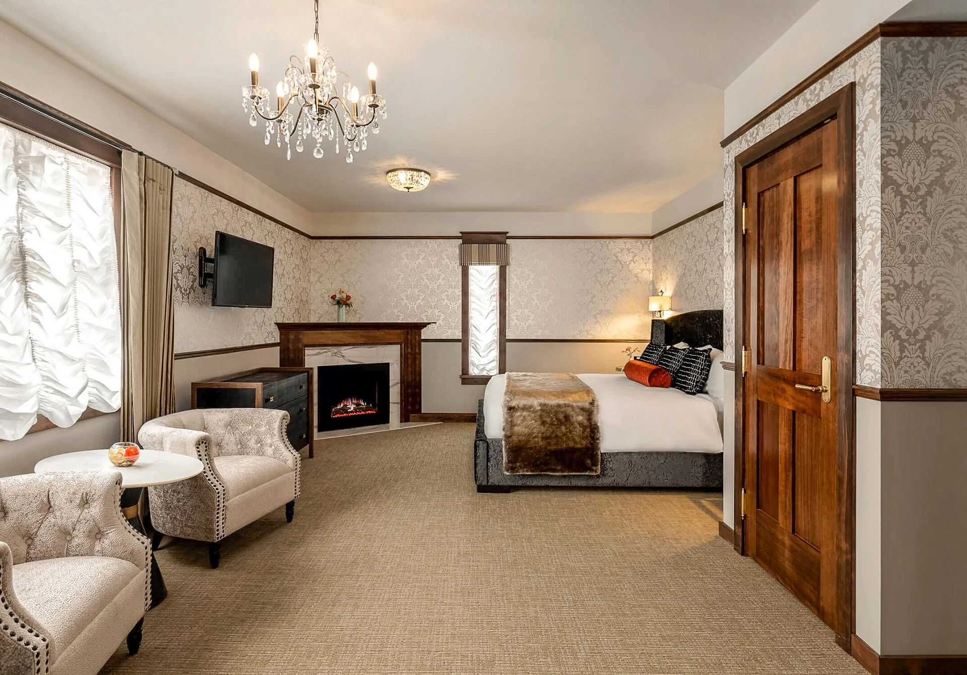 A spacious hotel bedroom with a bed, fireplace, wall-mounted TV, sitting area, chandelier, and elegant decor.