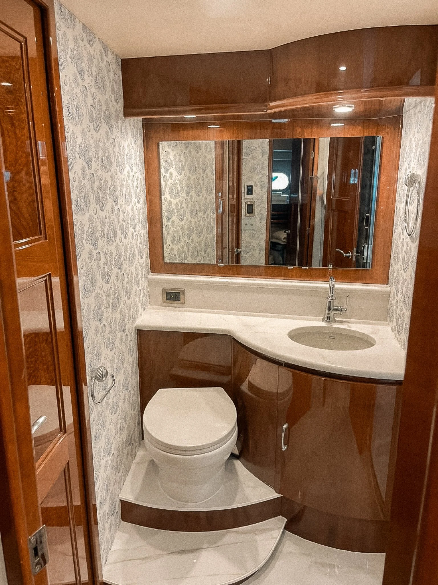 Small bathroom with a toilet, a marble countertop with a sink, a large mirror, wood cabinetry, and patterned wallpaper.