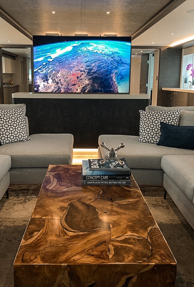 Living room with a large TV displaying Earth from space, two light gray sofas with patterned and dark pillows, a wooden coffee table with books and a decorative item, and a kitchen area in the background.