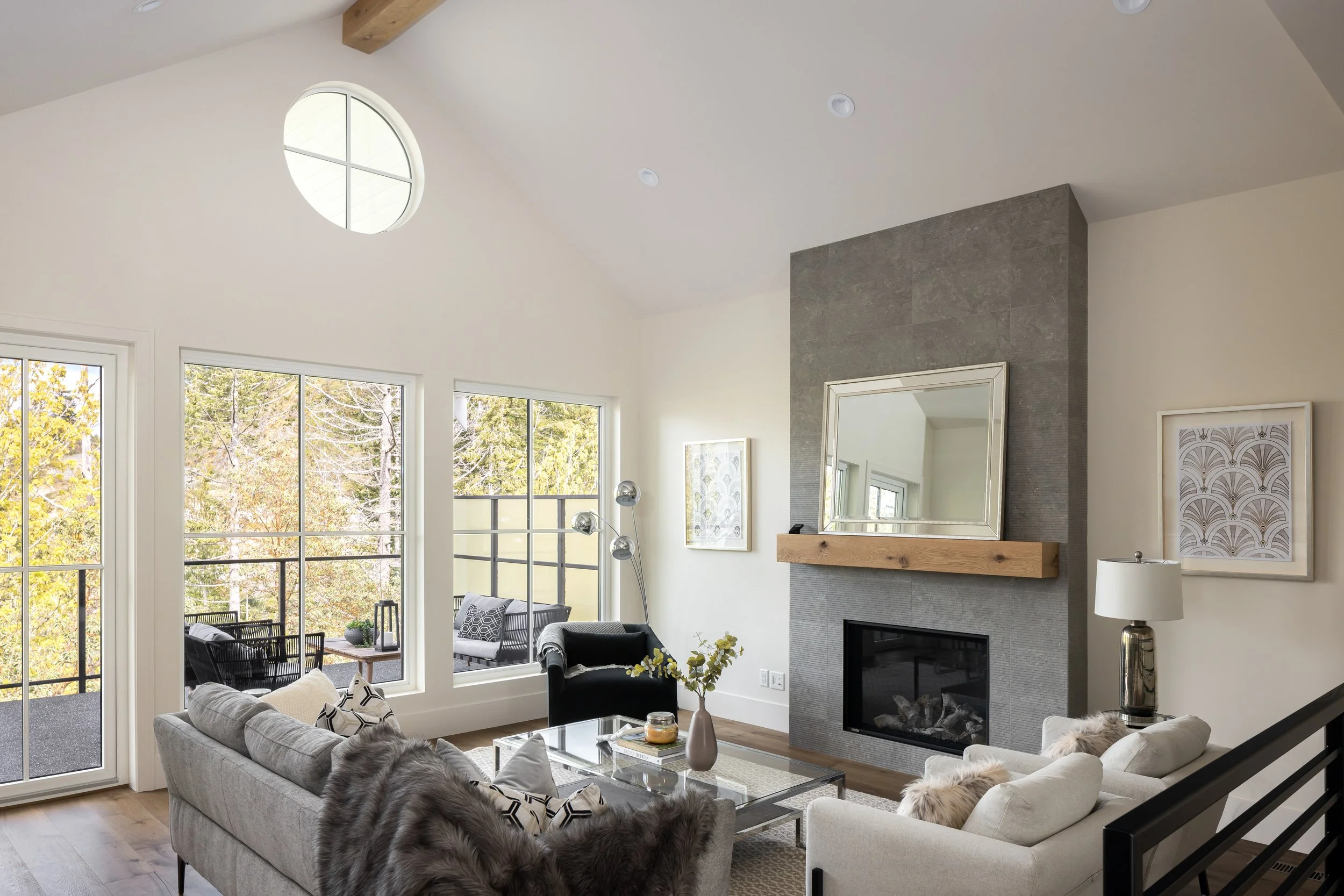 Living room with large windows, fireplace with a mirror and artwork, beige and black furniture, and views of a balcony with outdoor seating.