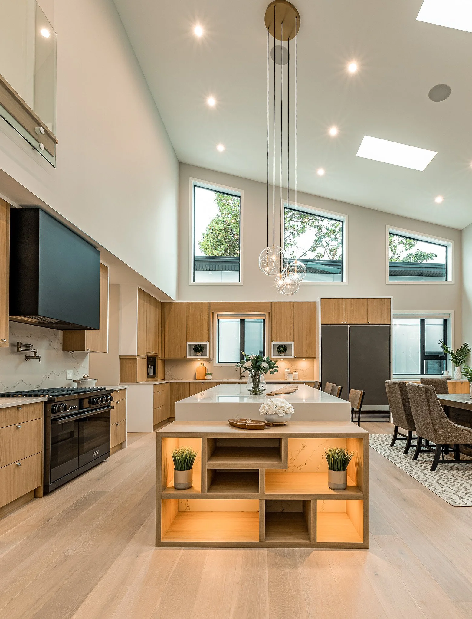 Open concept kitchen and dining area with high ceiling, large windows, and modern lighting fixture with glass bulbs.