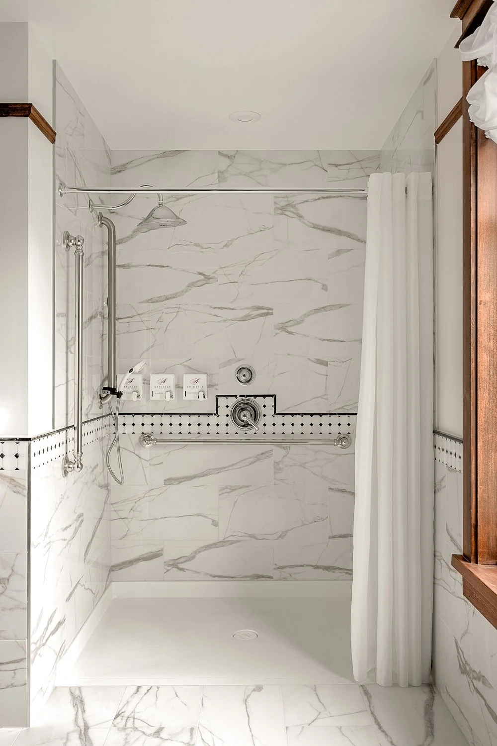 Empty shower with white marble walls with gray veins, a white curtain, and two shelves with toiletry bottles.