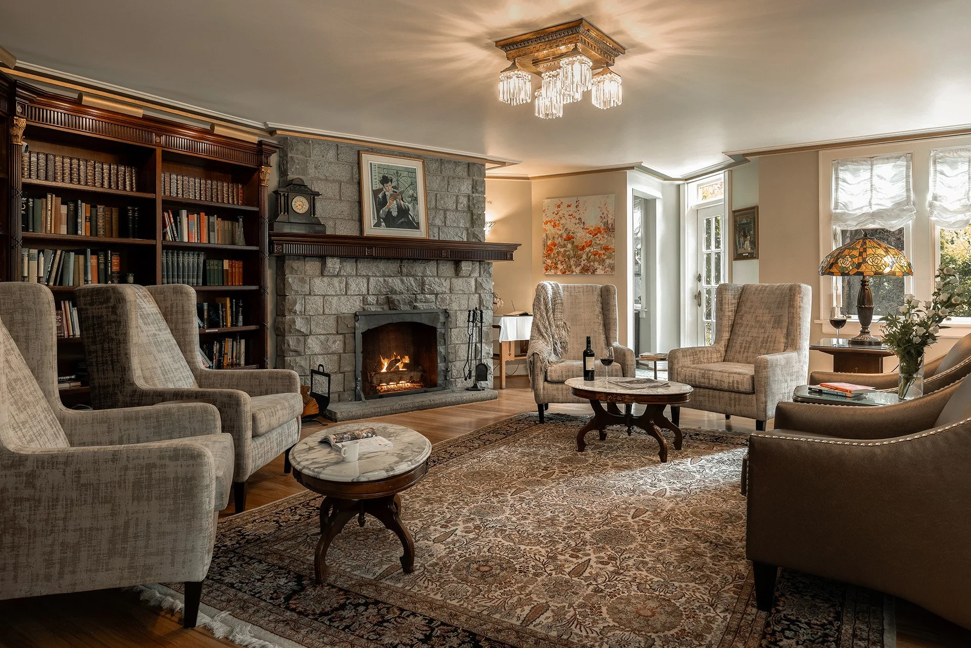 Cozy living room with a stone fireplace, armchairs, a patterned rug, a wooden coffee table with a bottle of wine and glasses, a book and teacup, and a large wooden bookshelf, decorated with picture frames, lamps, and artwork.