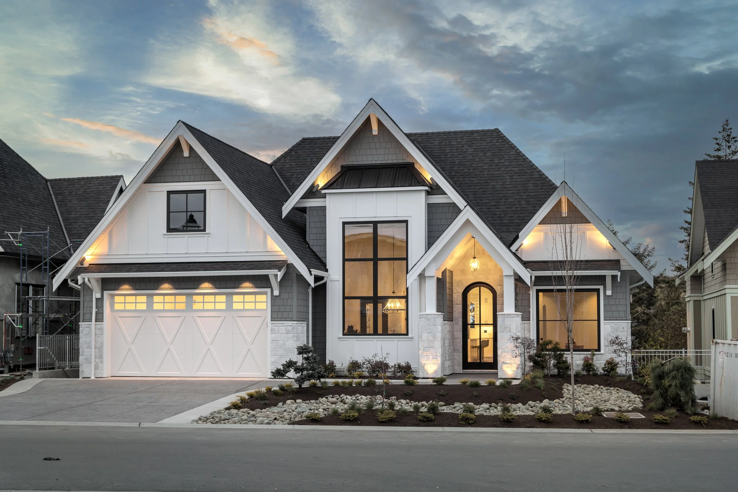 A modern two-story house with a dark shingled roof, white and gray exterior, large windows, and an illuminated front entrance, with landscaped yard and neighboring houses at dusk.