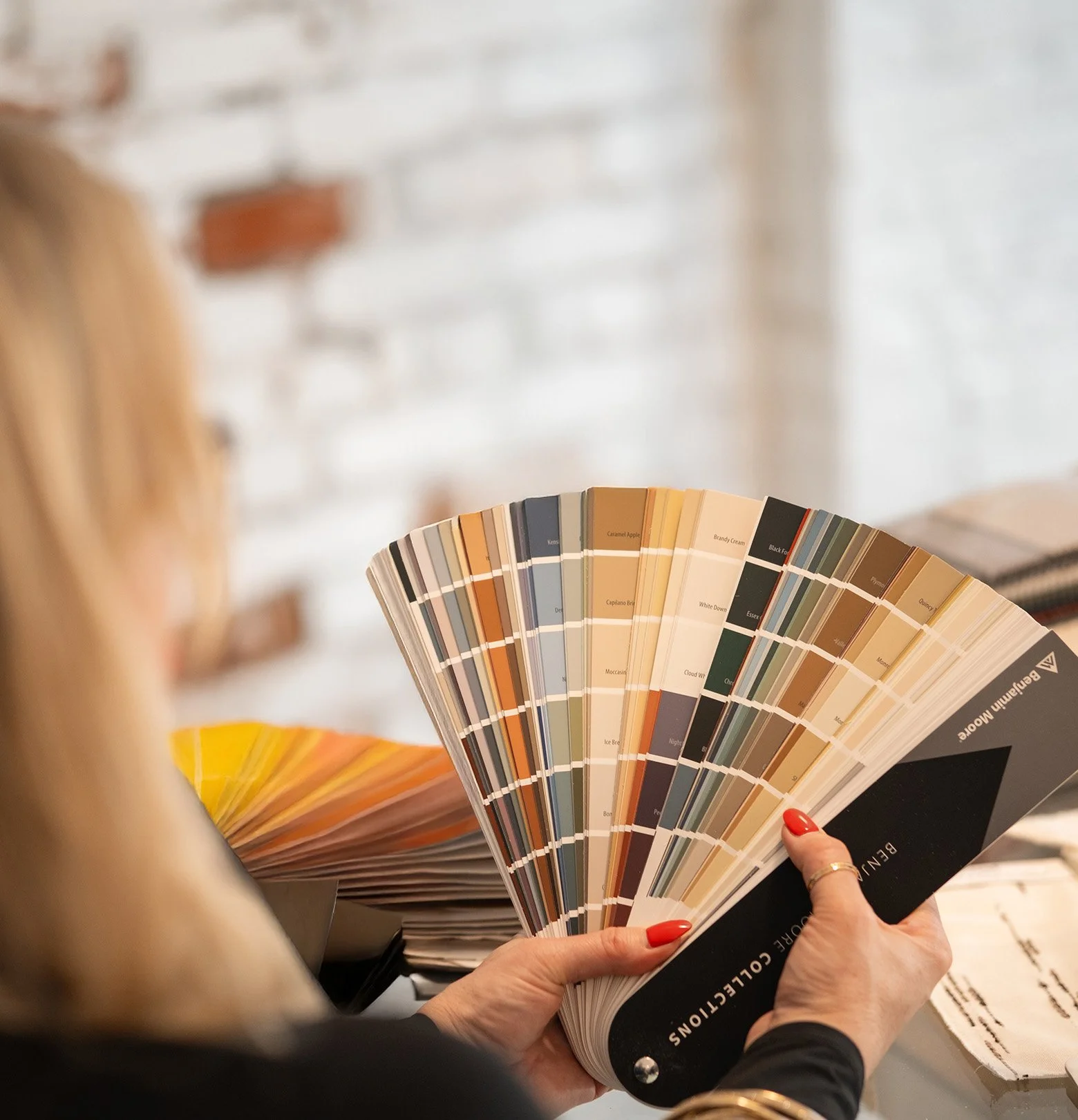 A person holding a fan-like paint color swatch book in a室 with a white brick wall background.