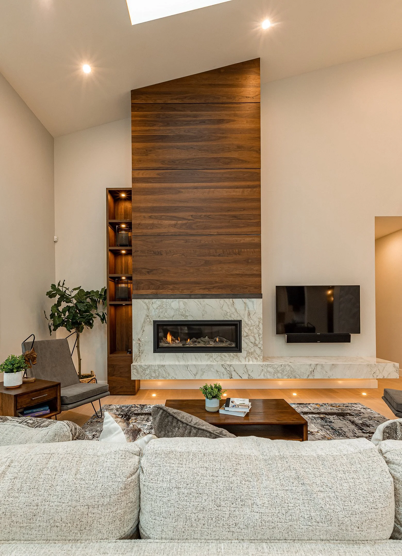 Modern living room featuring a fireplace with a marble surround, a mounted flat-screen TV, a beige couch, a wooden coffee table, and potted plants, with a high ceiling and built-in shelves.