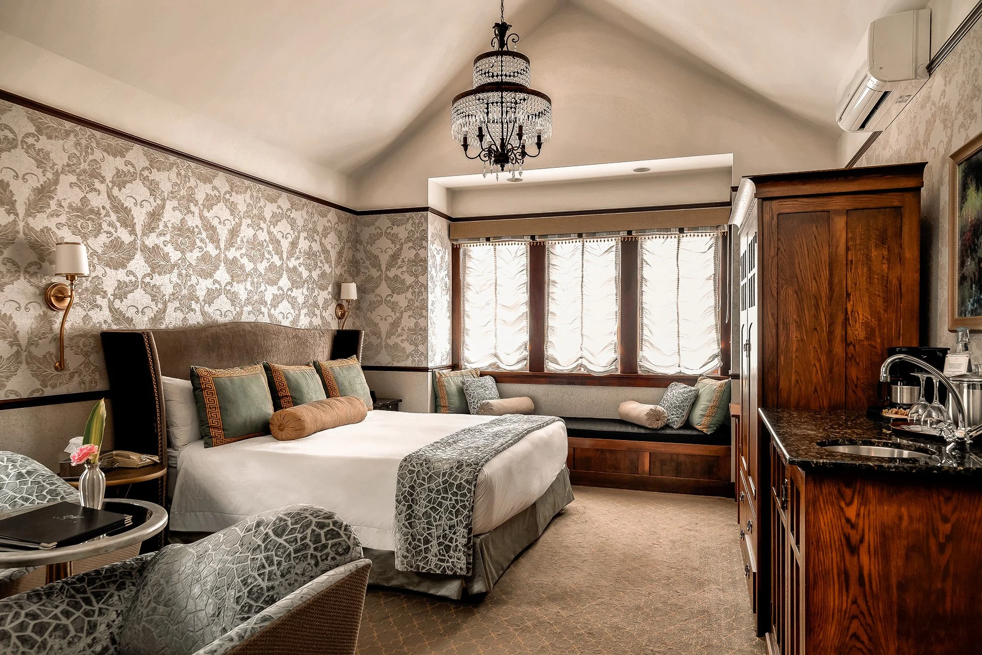 A luxurious hotel room featuring a large bed with decorative pillows, a cozy window seat with cushions, ornate wallpaper, a chandelier, a wooden cabinet, and a mini kitchenette, all illuminated by natural light from the window.