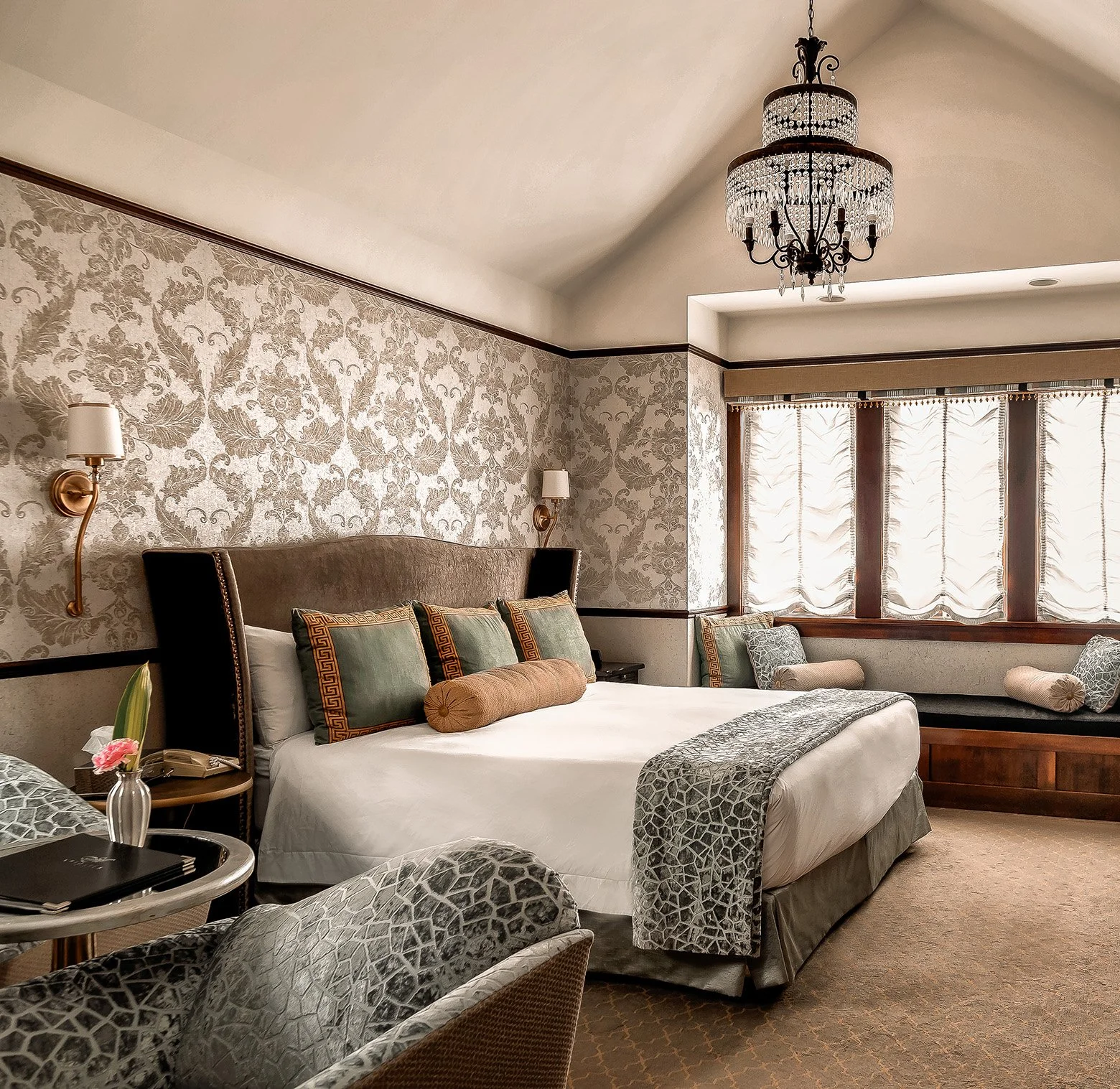 Luxurious hotel bedroom with a large bed, decorative pillows, a floral wallpaper, a chandelier, wall-mounted lamps, and large windows with curtains.