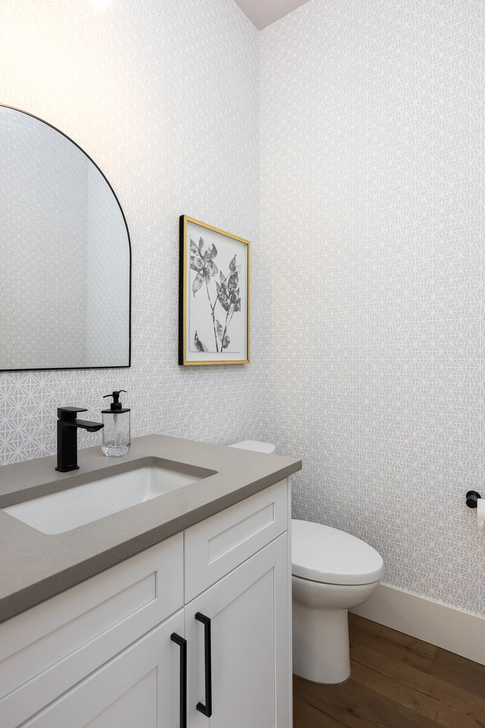 A modern bathroom featuring a white vanity with black handles, a beige countertop with a black faucet, a toilet, a framed black and white floral artwork, a large arched mirror, and geometric patterned wallpaper.