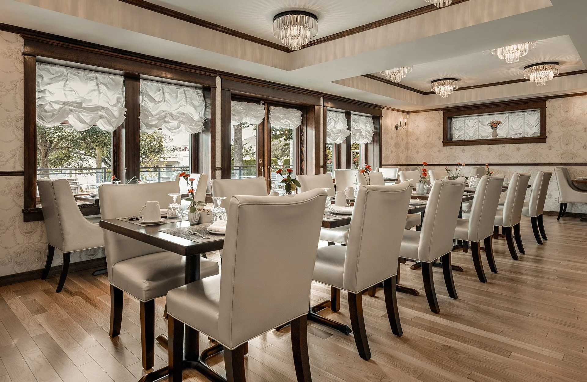Elegant dining room with white upholstered chairs arranged around dark wooden tables, decorated with small vases of orange flowers. Large windows with white curtains offer a view outside, and the room features a wooden floor, wallpaper, and ceiling chandeliers.
