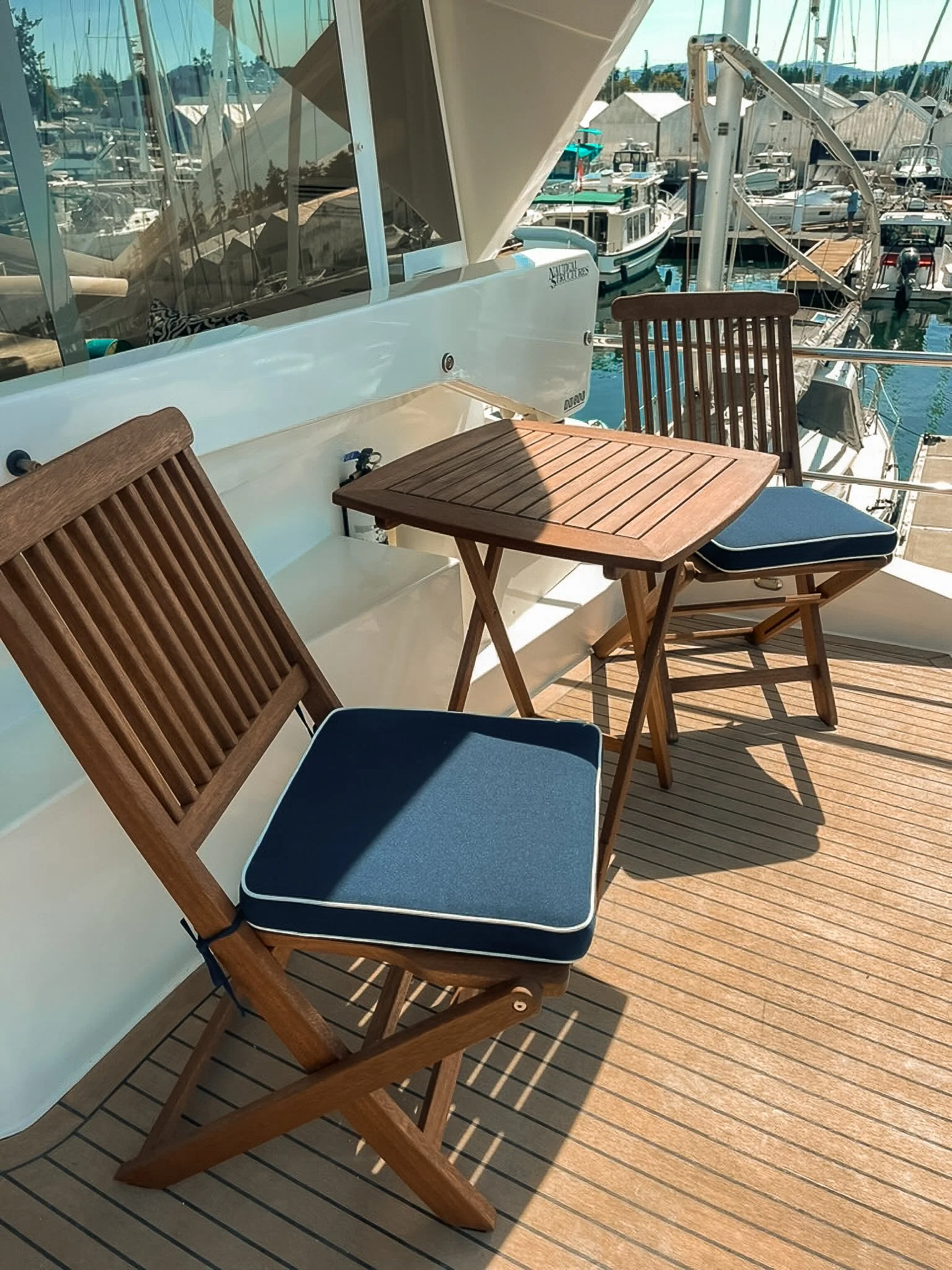 Two wooden chairs with blue cushions and a small wooden table on a boat deck, with a marina and boats in the background.