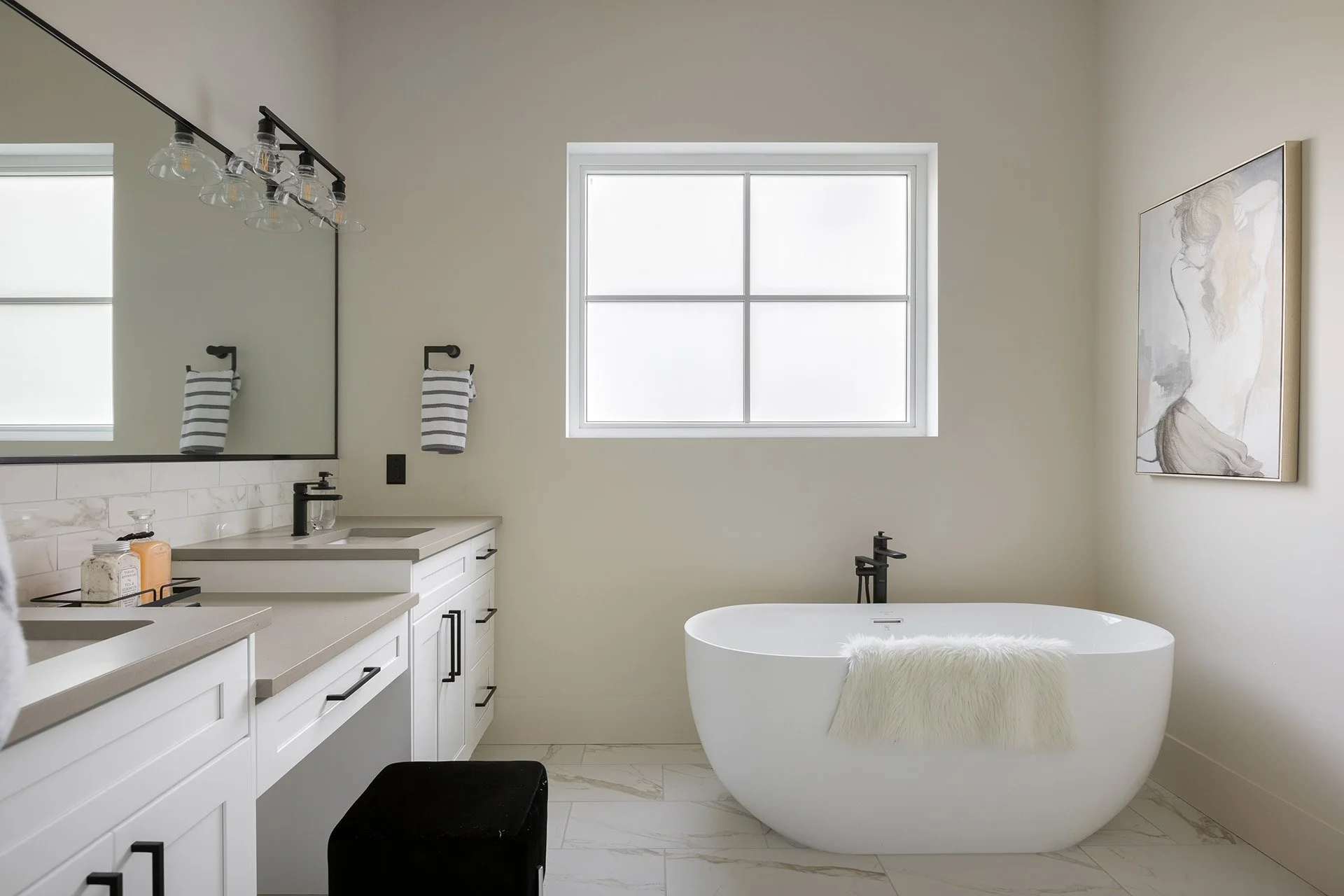 Modern bathroom with a white freestanding bathtub, a large mirror above white vanities, black fixtures, a window, and a wall art of a woman with long hair.