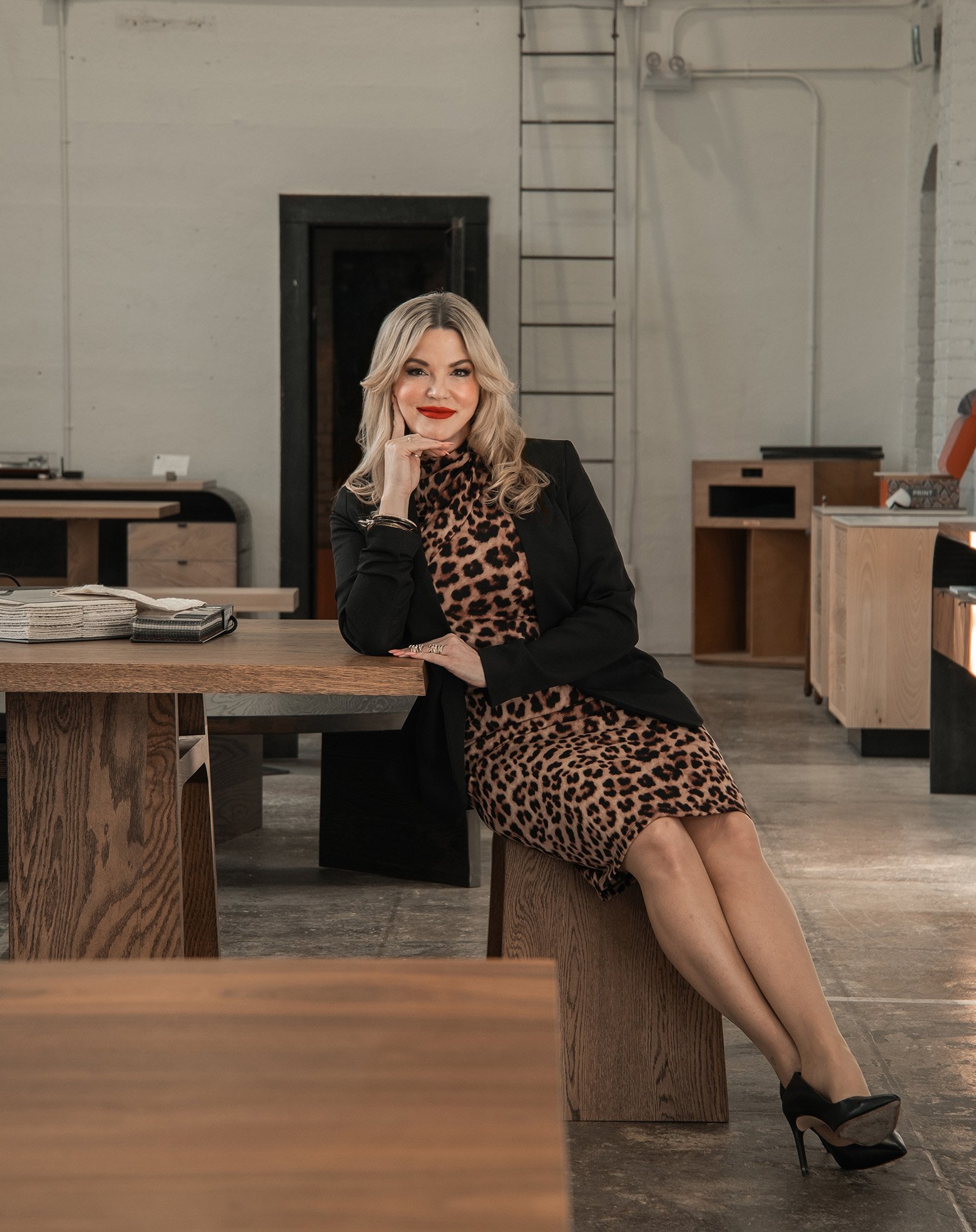 Danisha, a woman with blonde hair and red lipstick, sits on a bench at a wooden desk in an industrial-style office space, wearing a leopard print dress, black blazer, and heels.