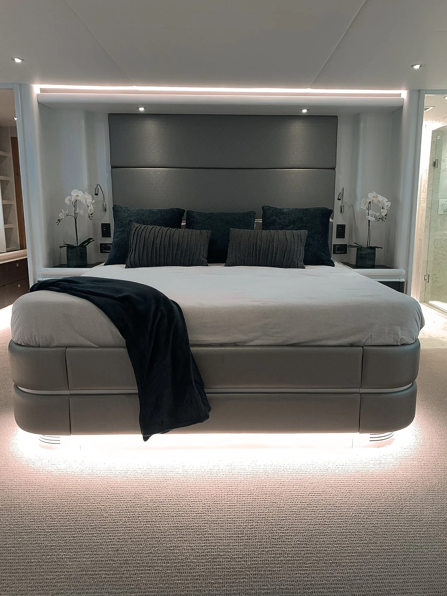Modern bedroom with a large bed, black and gray pillows, white bedding, black throw blanket, and orchid lamps on nightstands, illuminated by recessed lighting.