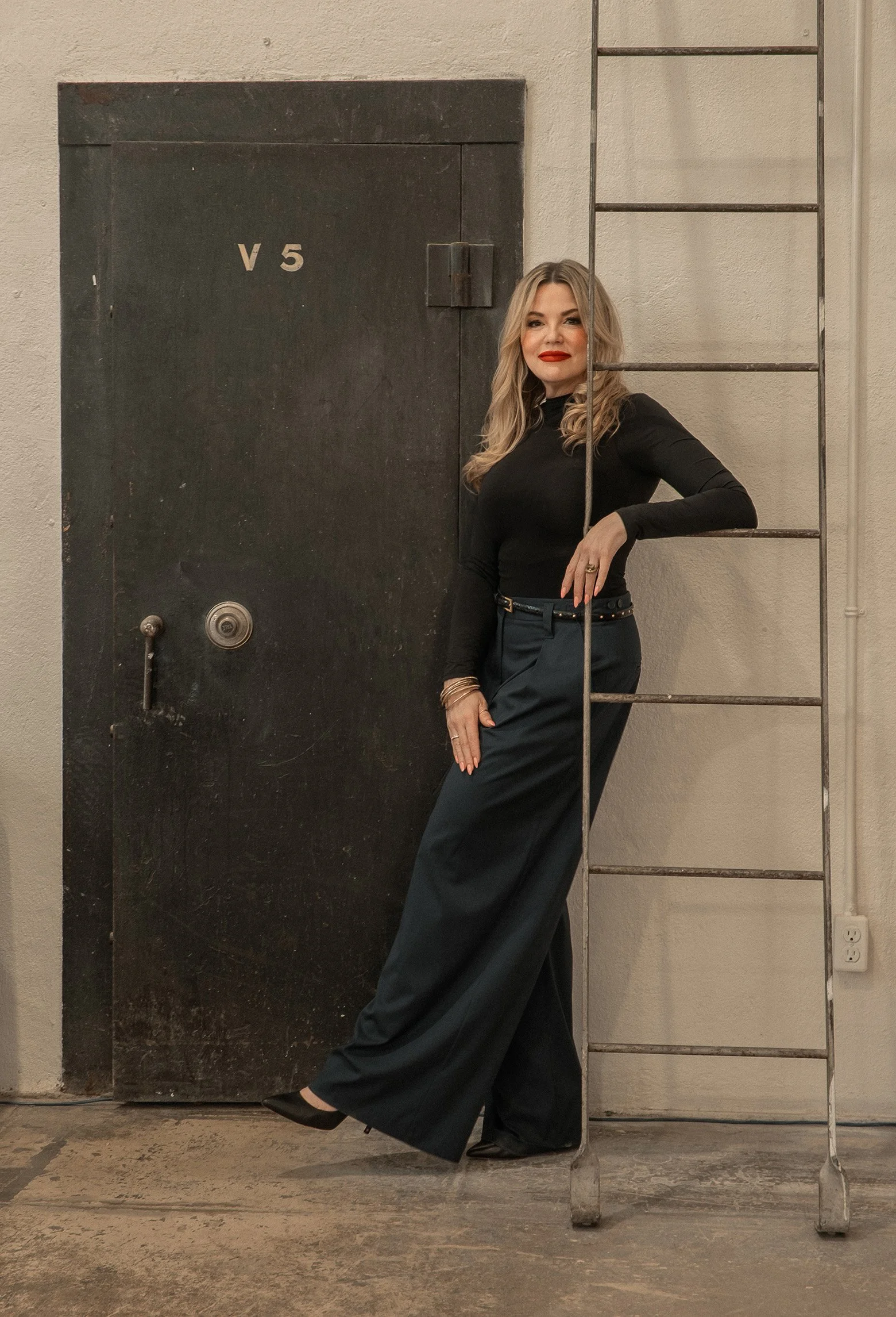 Danisha, a woman with blonde hair and red lipstick, stands against a grey wall, wearing a black long-sleeve top and wide-leg black pants, leaning on a metal ladder.