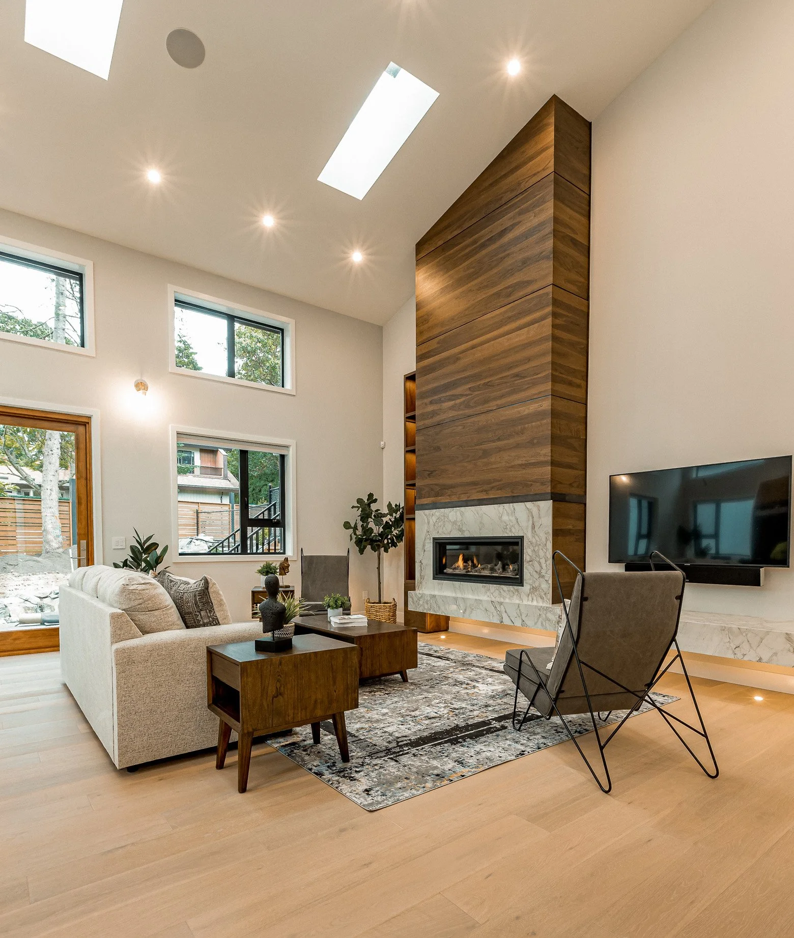 Modern living room with a beige sofa, a black lounge chair, a wooden coffee table, a fireplace with a marble surround, a mounted flat-screen TV, and large windows with natural light.