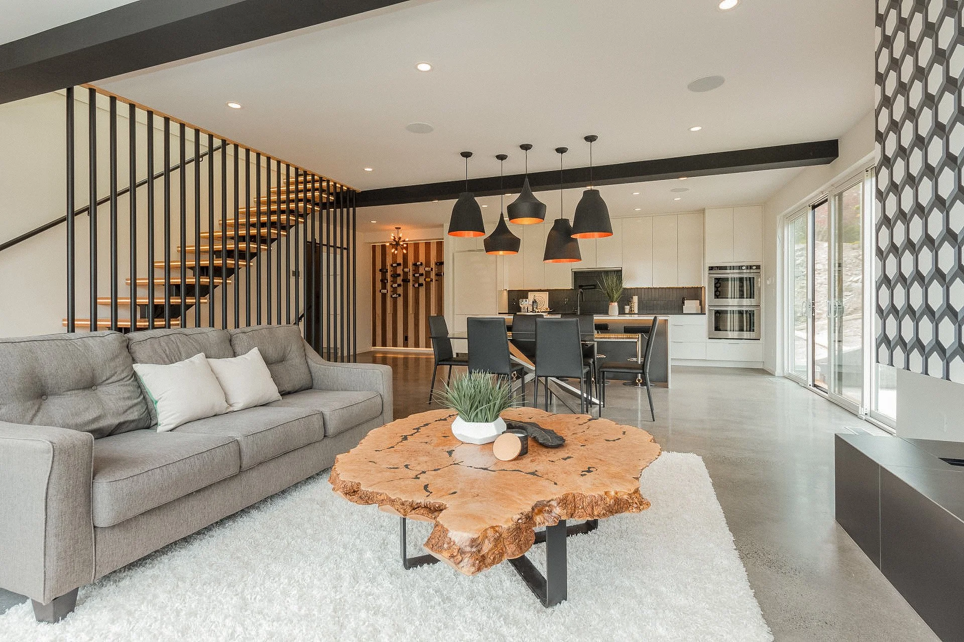 Open-concept living room and kitchen with modern decor, hardwood floors, a beige sofa with white pillows, a rustic wooden coffee table, black pendant lights over a dining table, white cabinetry, and large sliding glass doors leading outside.
