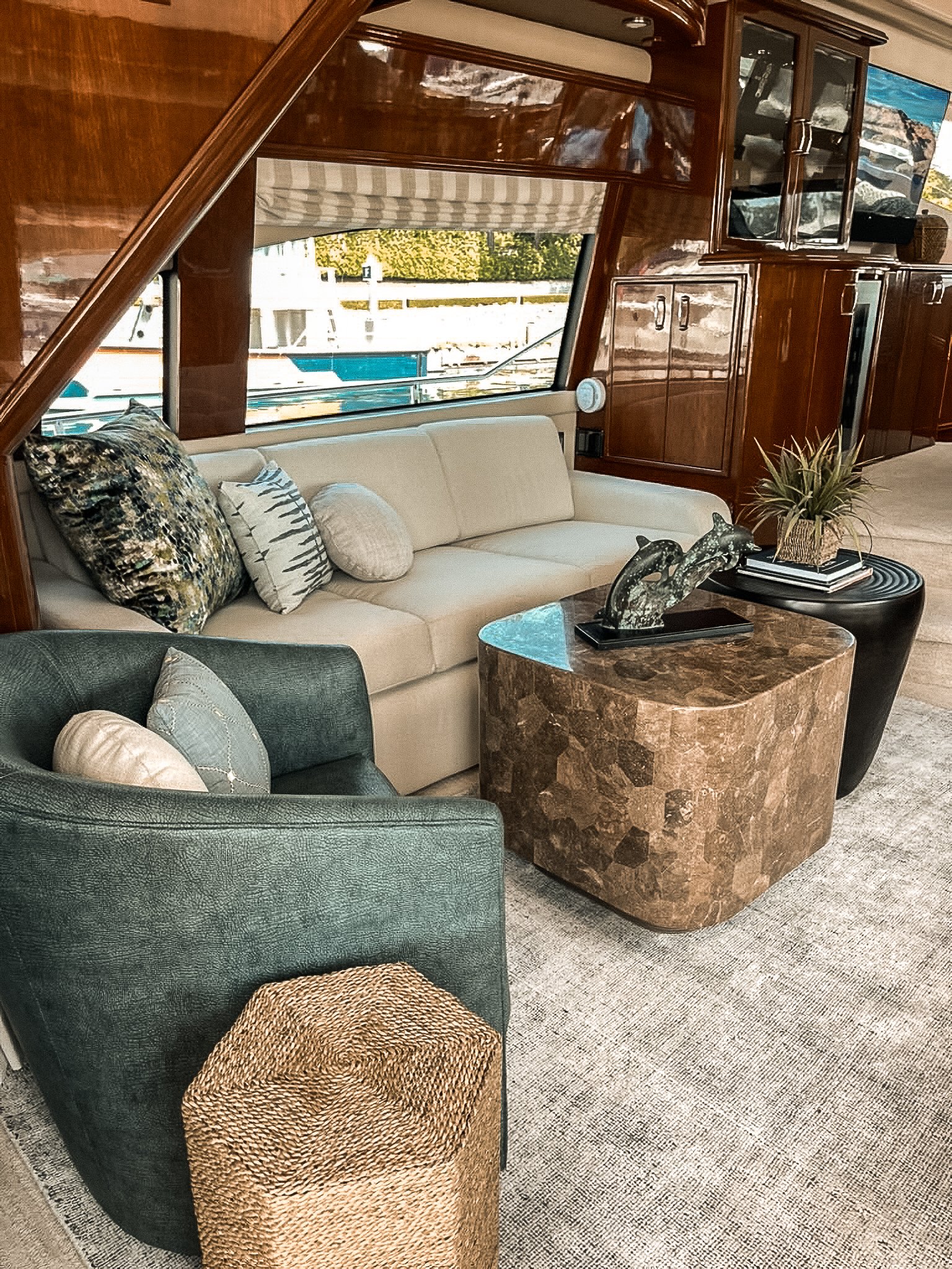 Interior of a boat with a cream sofa, dark green armchair, and marble coffee table in front of a large window showing boats docked outside. The decor includes patterned pillows, a potted plant, and sculptures on the table.