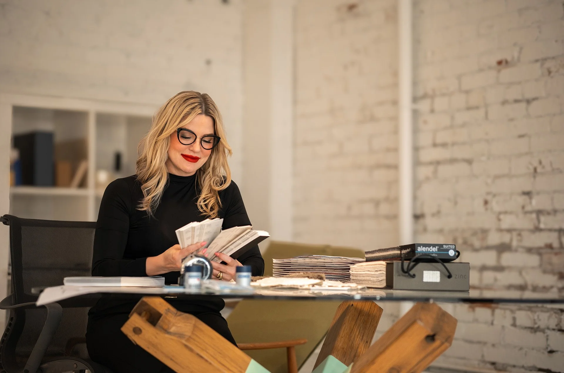 Danisha, a woman with glasses and blonde hair, sits at a desk, reading a booklet, surrounded by papers and office supplies in a modern workspace.