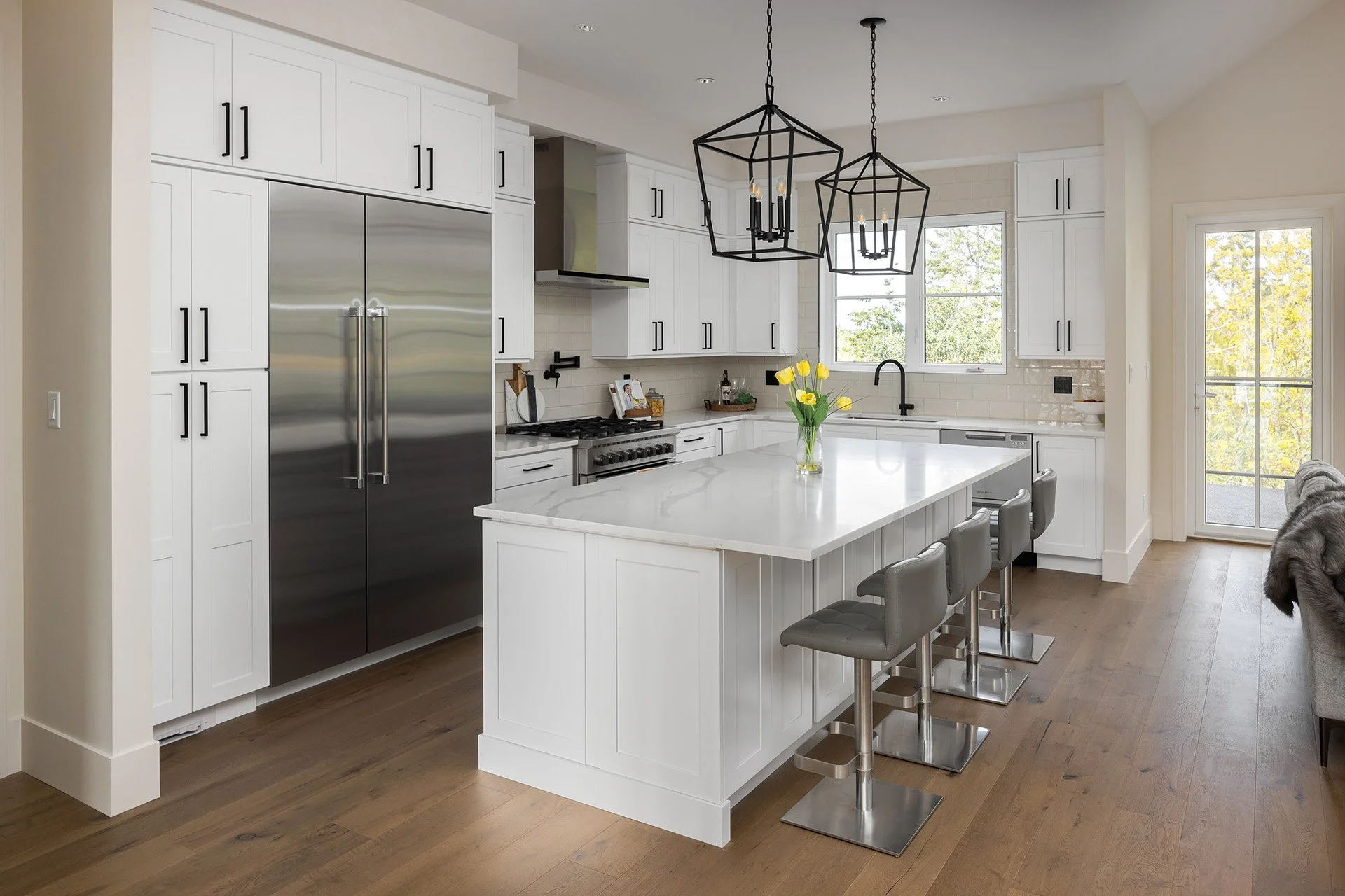 Modern kitchen with white cabinetry, stainless steel appliances, a kitchen island with a white countertop, four grey bar stools, black pendant lights, a window with a view of trees, and yellow tulips in a glass vase on the island.