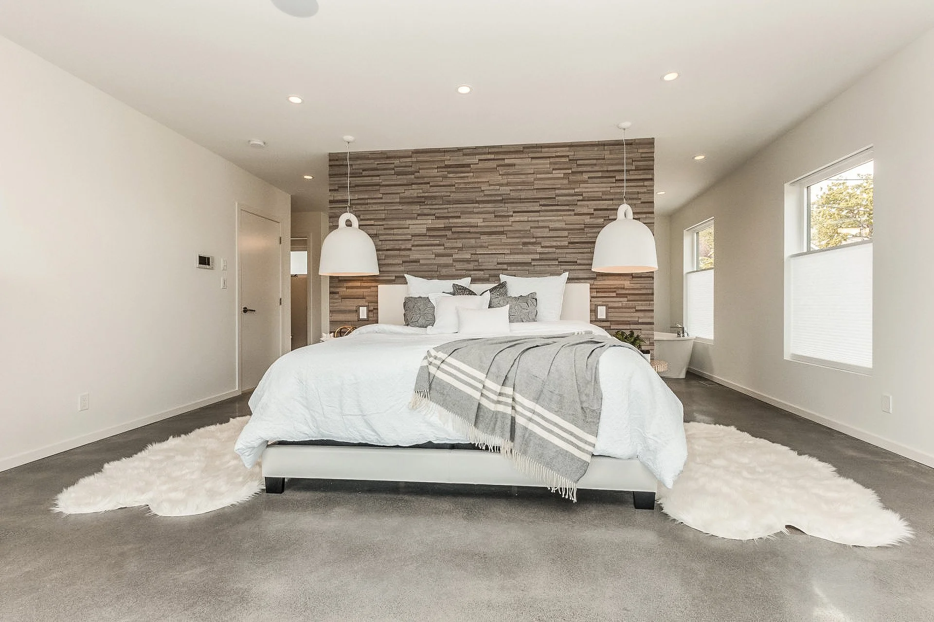 Modern bedroom with a white bed, gray throw blanket, hardwood headboard wall, hanging pendant lamps, and fluffy white rugs on gray carpet.