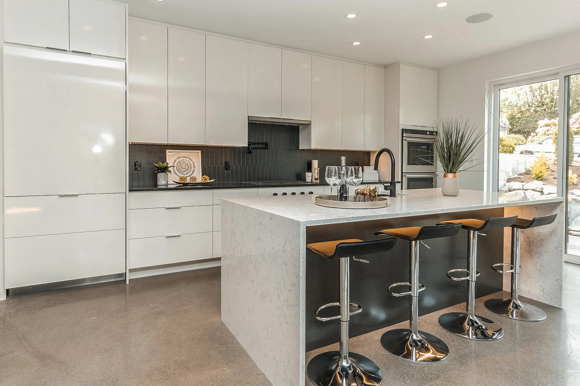 Modern kitchen with white cabinetry, black backsplash, and a large white marble island with four black and brown bar stools. Decor includes a potted plant, wine glasses, and a tray with small figurines. Large window shows outdoor landscaping.